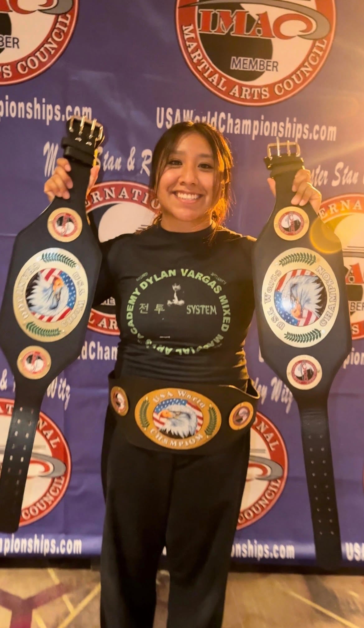 A smiling woman stands in front of a blue promotional backdrop holding two large martial arts championship belts, with a black graphic T-shirt and black pants visible. The backdrop includes USA World Championships branding and a TMAQ Martial Arts Council logo.