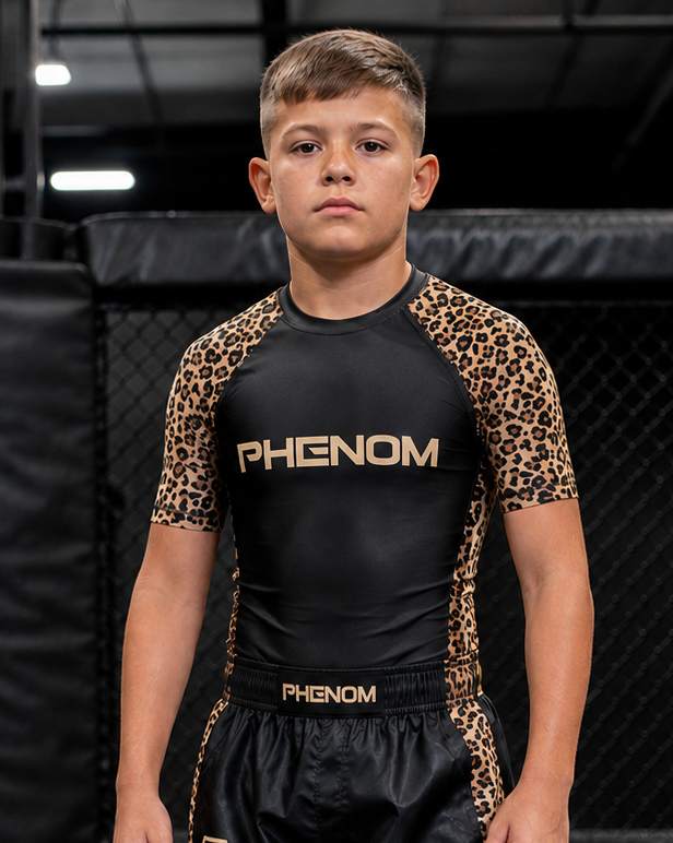 A young boy stands barefoot inside an MMA or boxing cage wearing a black rash guard with leopard print sleeves and side panels, and matching black shorts with gold branding.