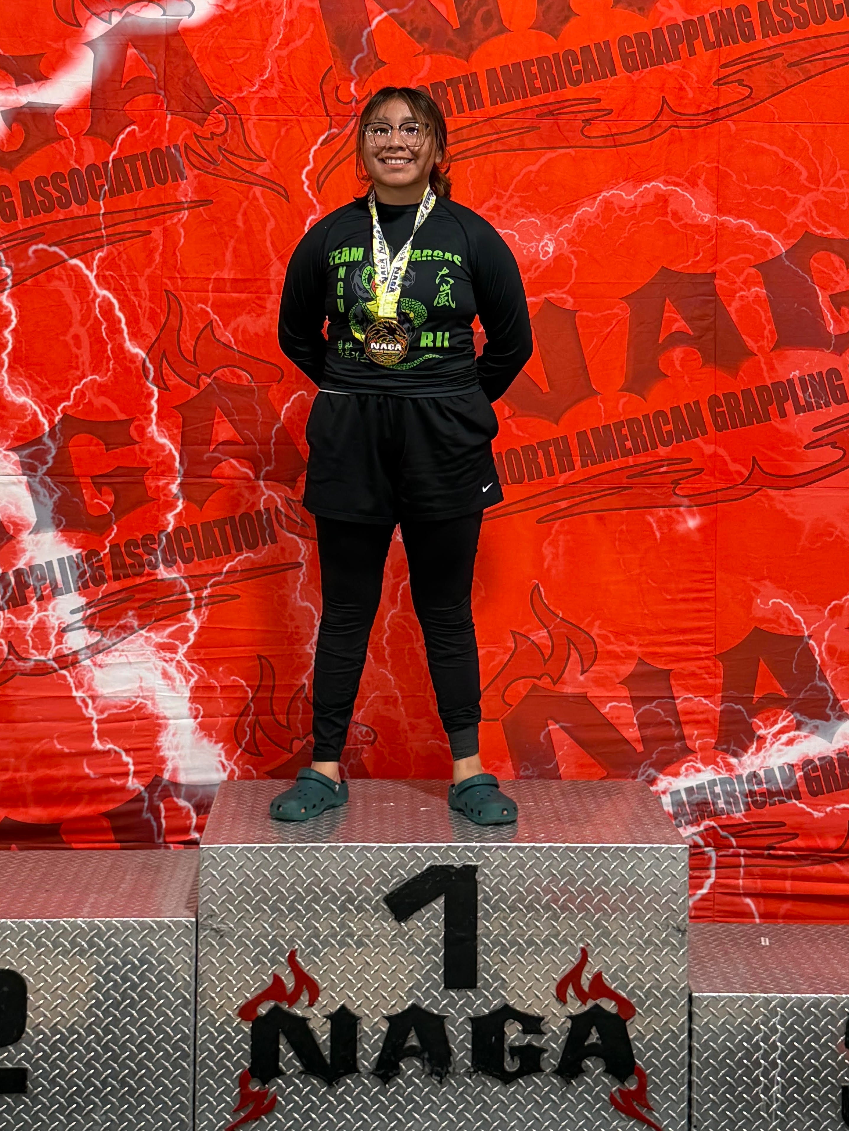 A woman stands on a 1st-place podium at a NAGA wrestling event, holding a medal and trophy in front of a large red and black NAGA backdrop.