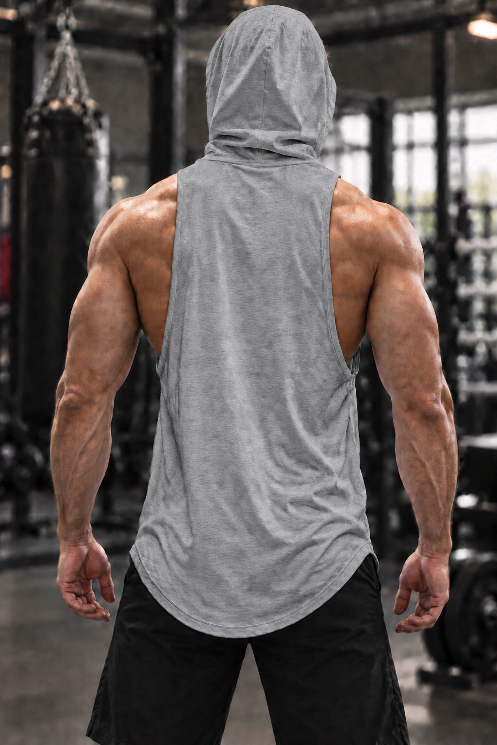 A muscular man viewed from behind wearing a heather gray sleeveless hooded tank top and black shorts in a gym setting.