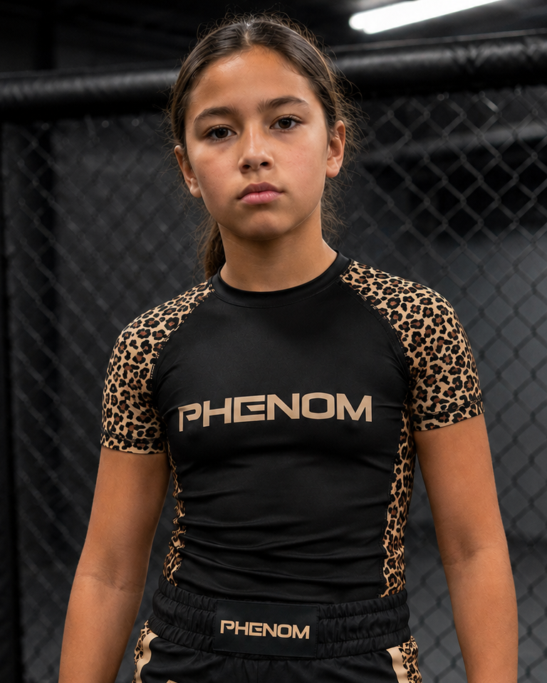 A young female athlete wearing a black and leopard-print Phenom rash guard and matching black shorts with gold trim, standing inside a black MMA cage.