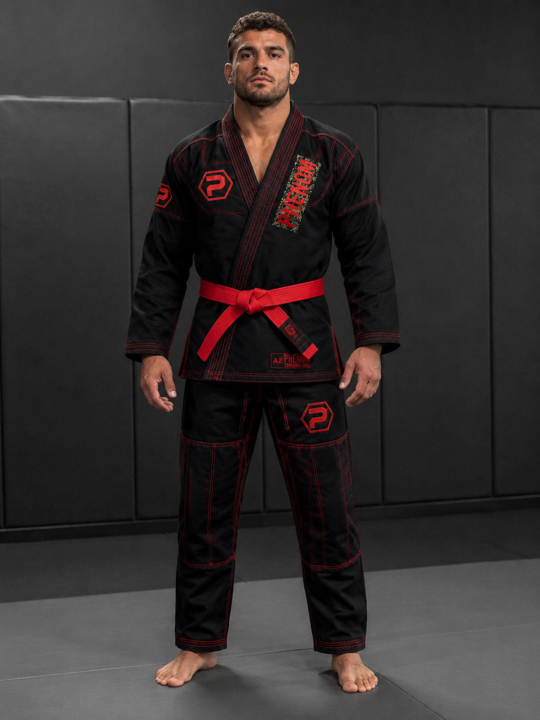 A man wearing a black Brazilian Jiu-Jitsu gi with red contrast stitching and a red belt, standing on a dark padded mat.