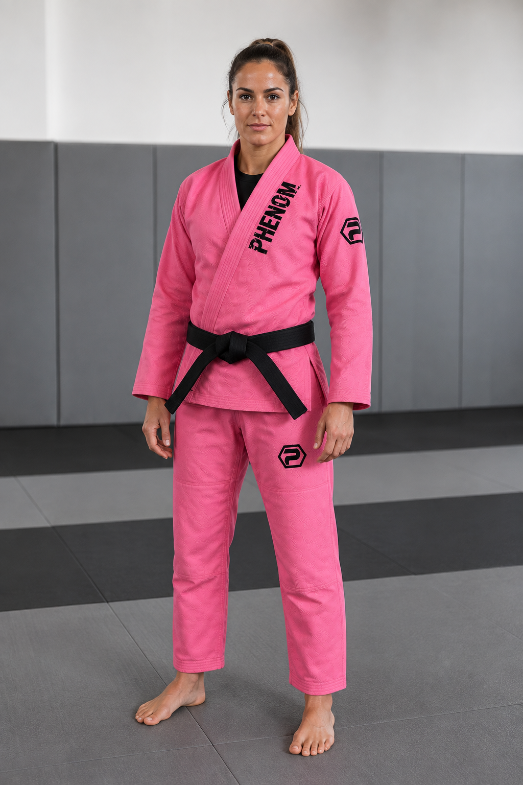 A woman wearing a bright pink Brazilian Jiu-Jitsu gi with a black belt and black printed branding on the chest and sleeve, standing in a gym with gray mats.