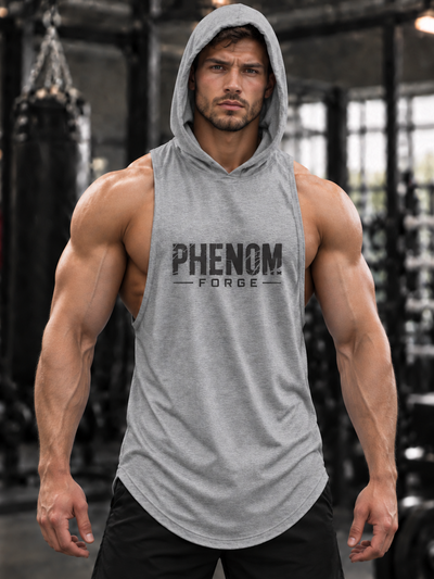 A muscular man wearing a heather gray sleeveless hooded tank top with 'PHENOM FORGE' printed in black across the chest, standing in a gym with a blurred punching bag in the background.