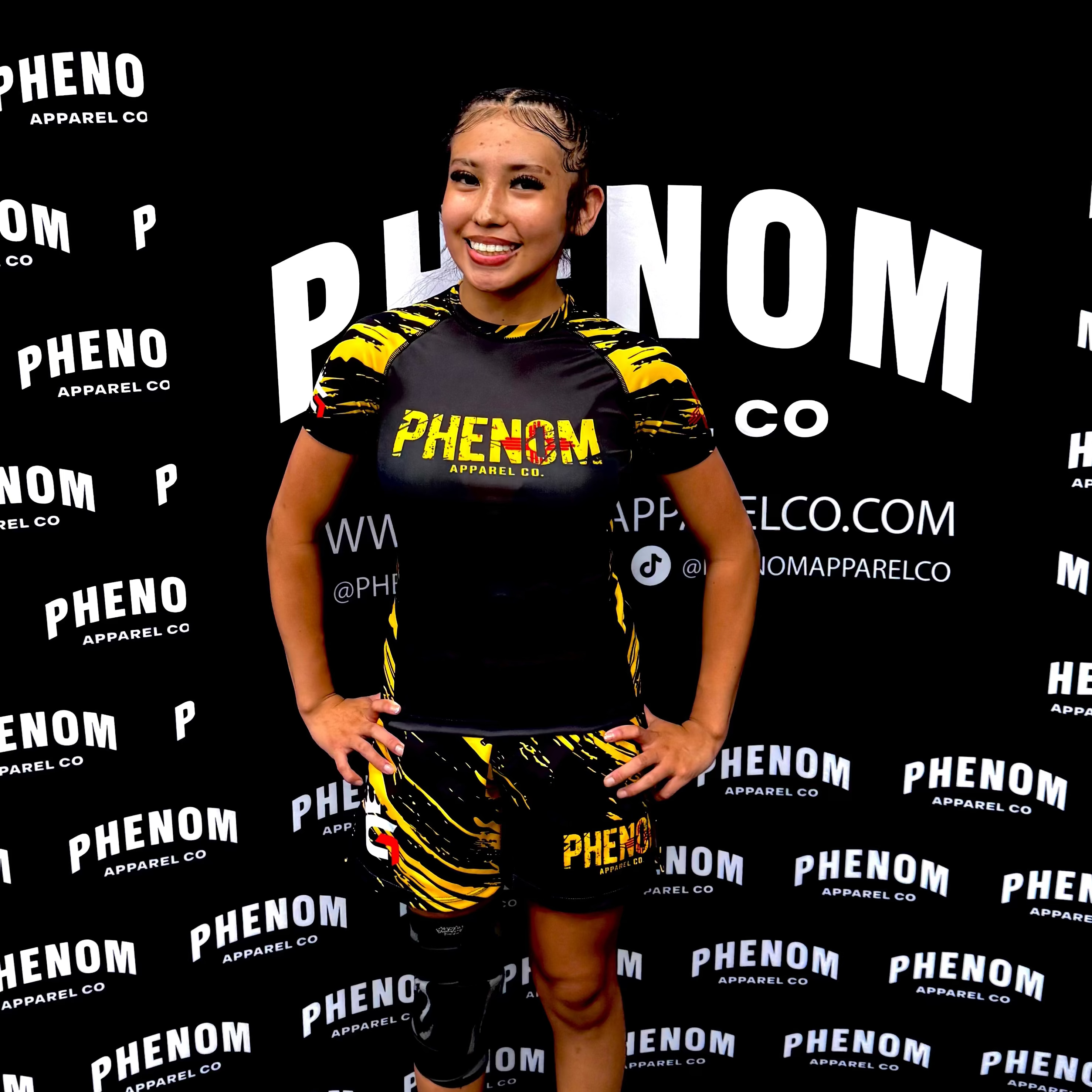 A FEMALE ATHLETE stands in front of a branded black backdrop wearing a black and yellow Phenom apparel set consisting of a short-sleeve top and matching shorts. The backdrop is covered in repeated Phenom logos and text, and the model is wearing black sandals.