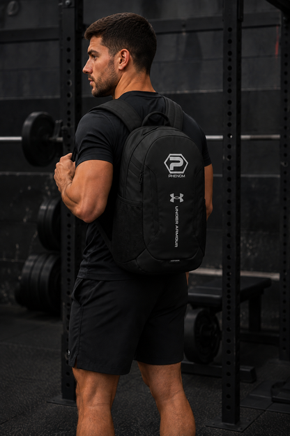 A man in a black athletic outfit seen from behind wearing a black Under Armour backpack with a prominent white phenom  logo on the back, set in a dark gym environment.