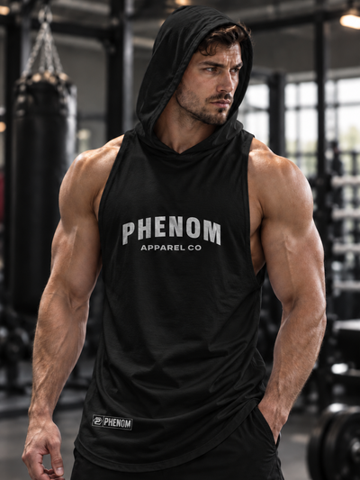 A muscular man in a gym wearing a black sleeveless hooded tank top with 'PHENOM APPAREL CO' printed in white on the chest, paired with black shorts.