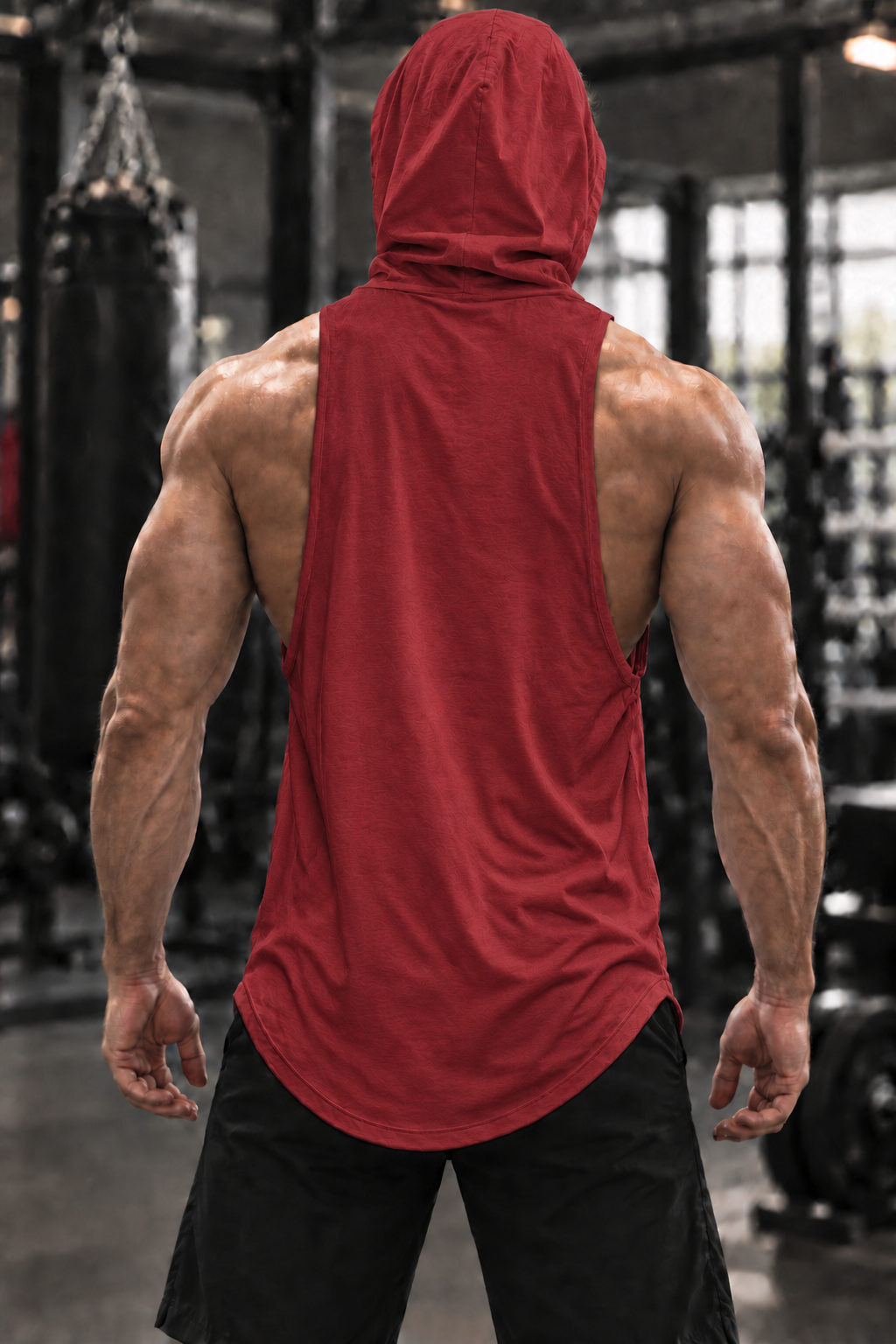 A muscular man viewed from behind wearing a red sleeveless hooded tank top and black shorts in a gym setting.