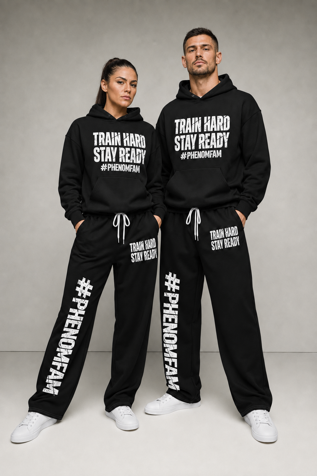 A man and a woman modeling matching black hoodies and sweatpants sets with white printed text reading 'TRAIN HARD STAY READY #PHENOMENAFAN' against a neutral gray studio background.