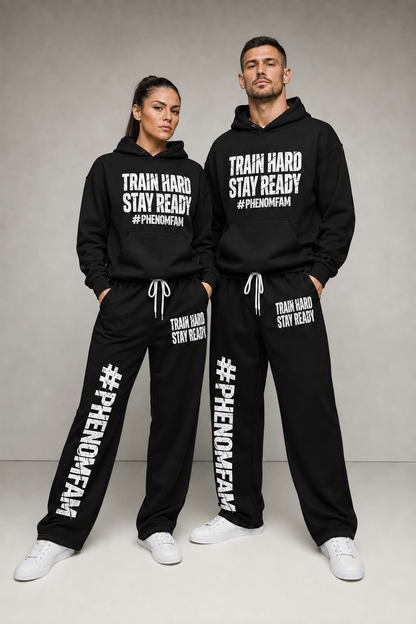 A man and a woman modeling matching black hoodies and sweatpants sets with white printed text reading 'TRAIN HARD STAY READY #PHENOMENAFAN' against a neutral gray studio background.