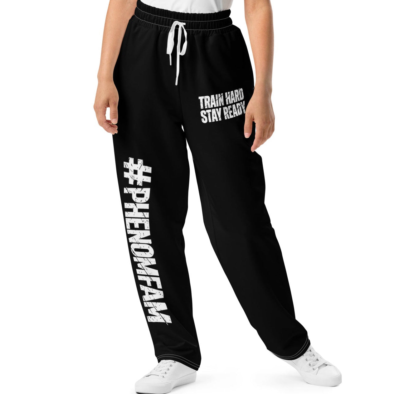 A woman modeling black sweatpants with white text and drawstrings, paired with a white tank top and white sneakers, against a plain white background.