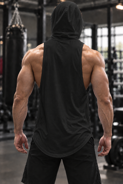 A muscular man viewed from behind wearing a black sleeveless hooded tank top and black shorts in a gym setting.