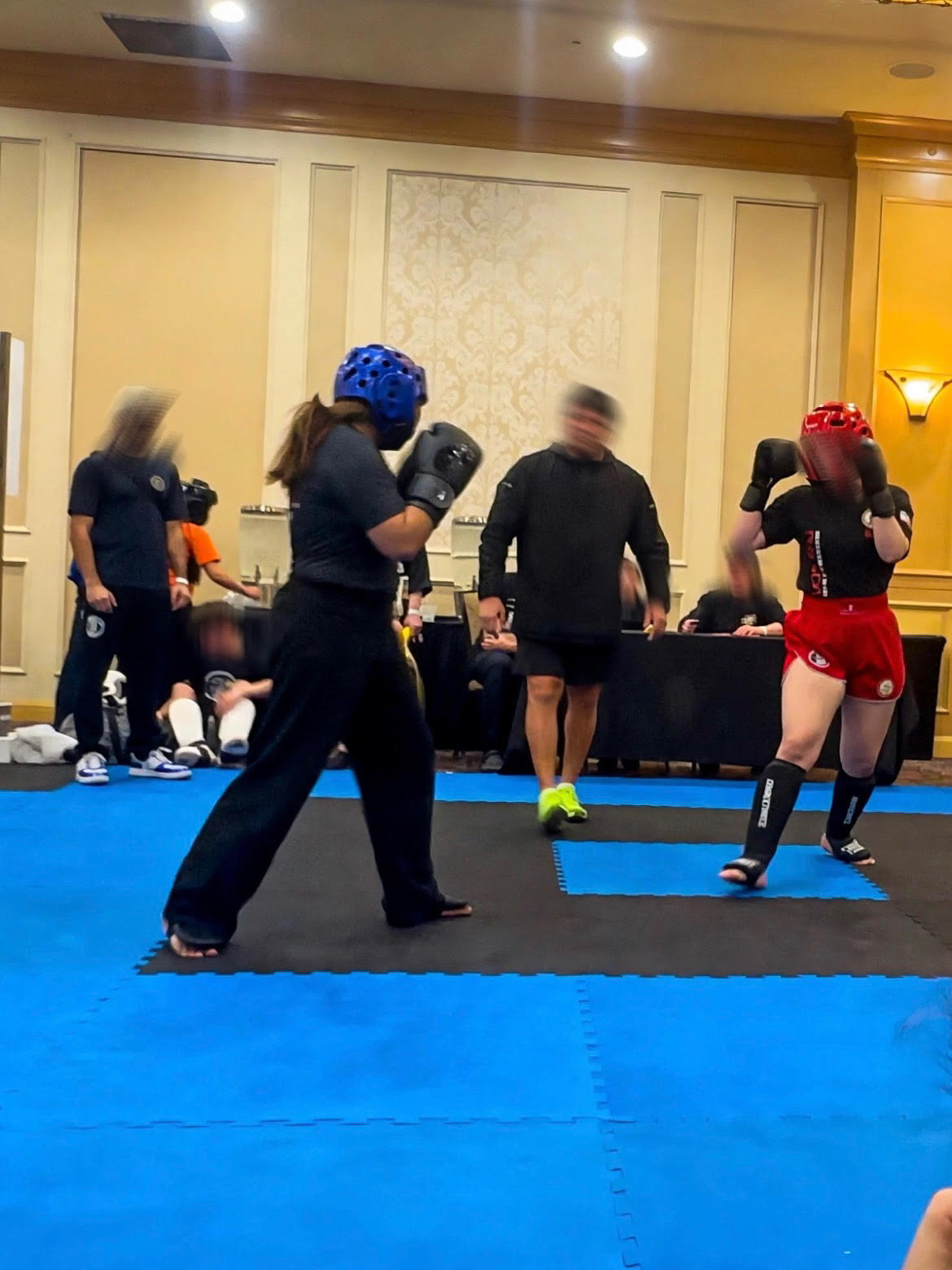 Two people in athletic gear sparring on blue and black mats inside a gym, with one person wearing a blue headguard and black outfit, and the other wearing a red headguard and red shorts.