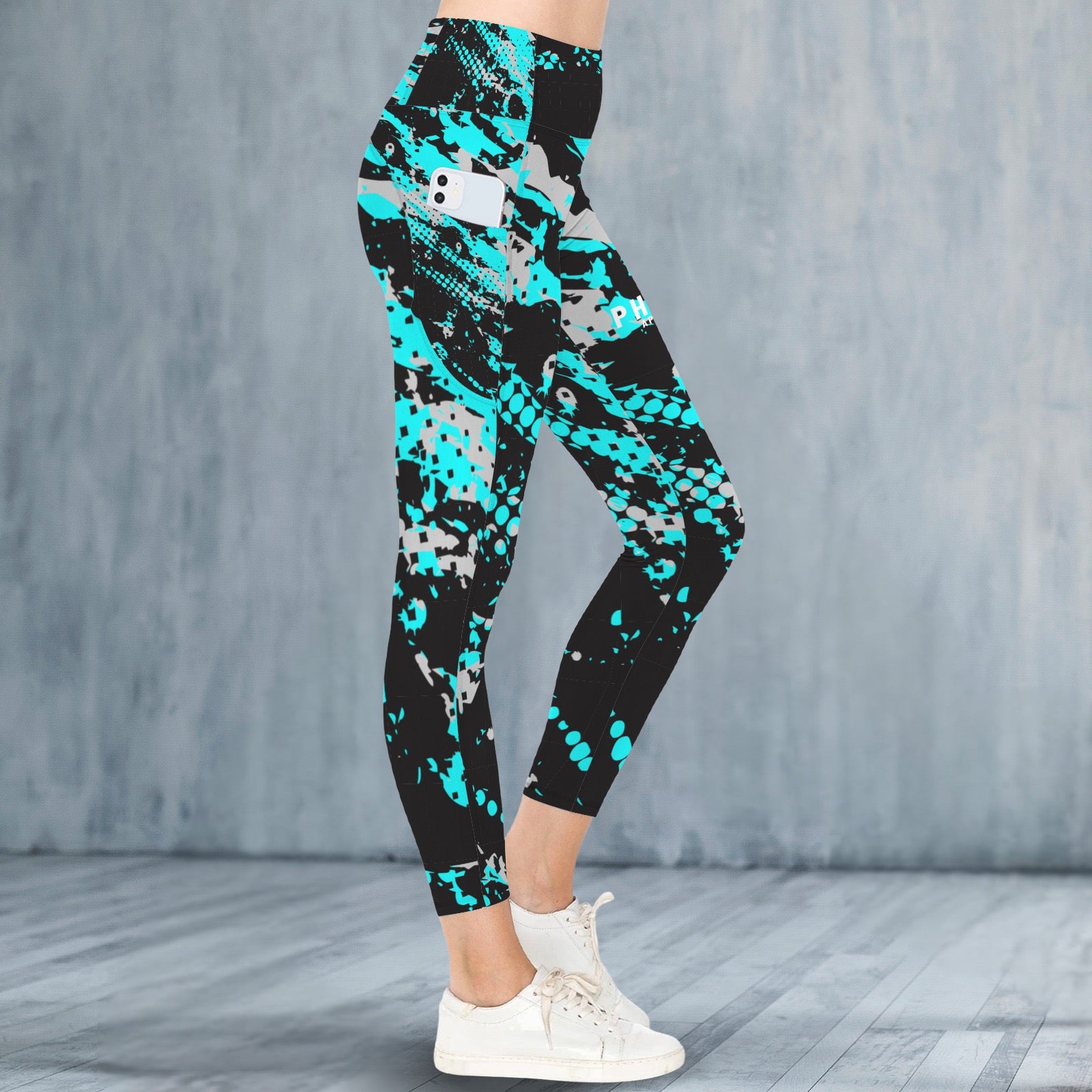 A person wearing black leggings with a bold turquoise, gray, and white abstract pattern, paired with white sneakers, standing against a textured gray wall and wood floor.