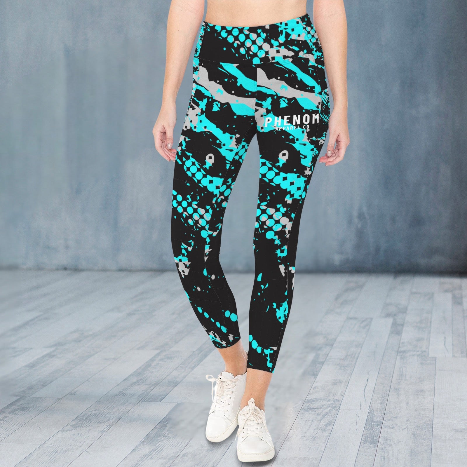 A model from the waist down wearing high-waisted athletic leggings with a black, teal, and gray shark-scale pattern, paired with white sneakers, set against a gray studio backdrop.