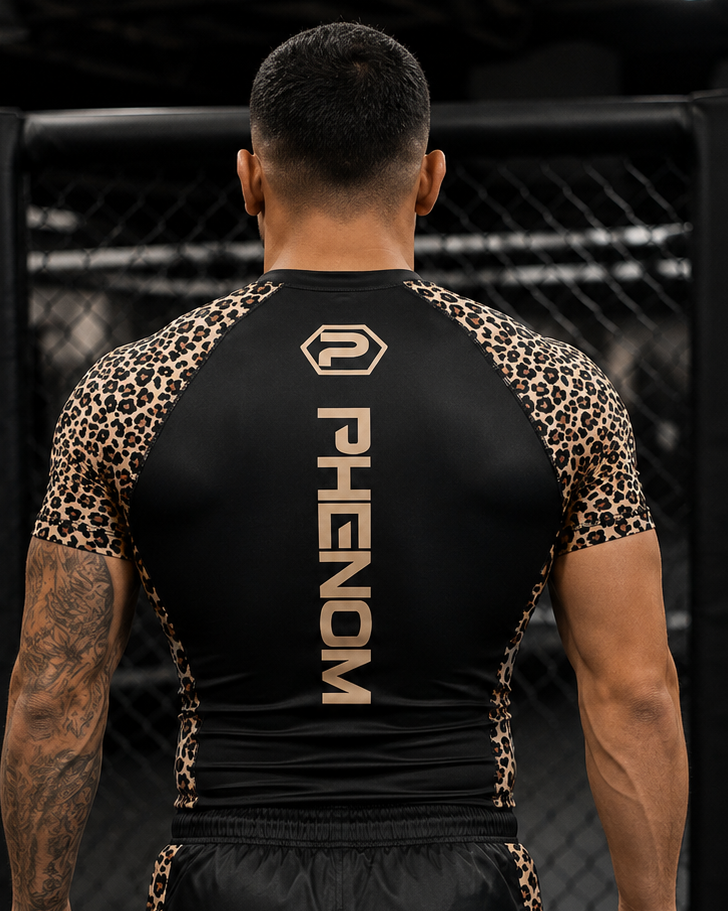A man viewed from behind wearing a black athletic rash guard with leopard-print sleeves and side panels, plus matching black shorts with leopard-print side stripes. The back of the rash guard is black with vertical PHENOM branding in a tan/gold tone.