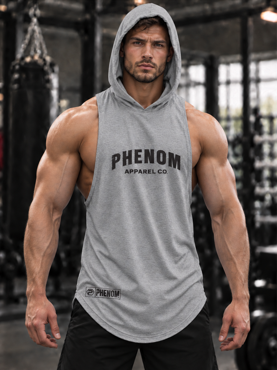 A muscular man wearing a heather gray sleeveless hooded tank top with 'PHENOM APPAREL CO' printed in black across the chest, standing in a gym.
