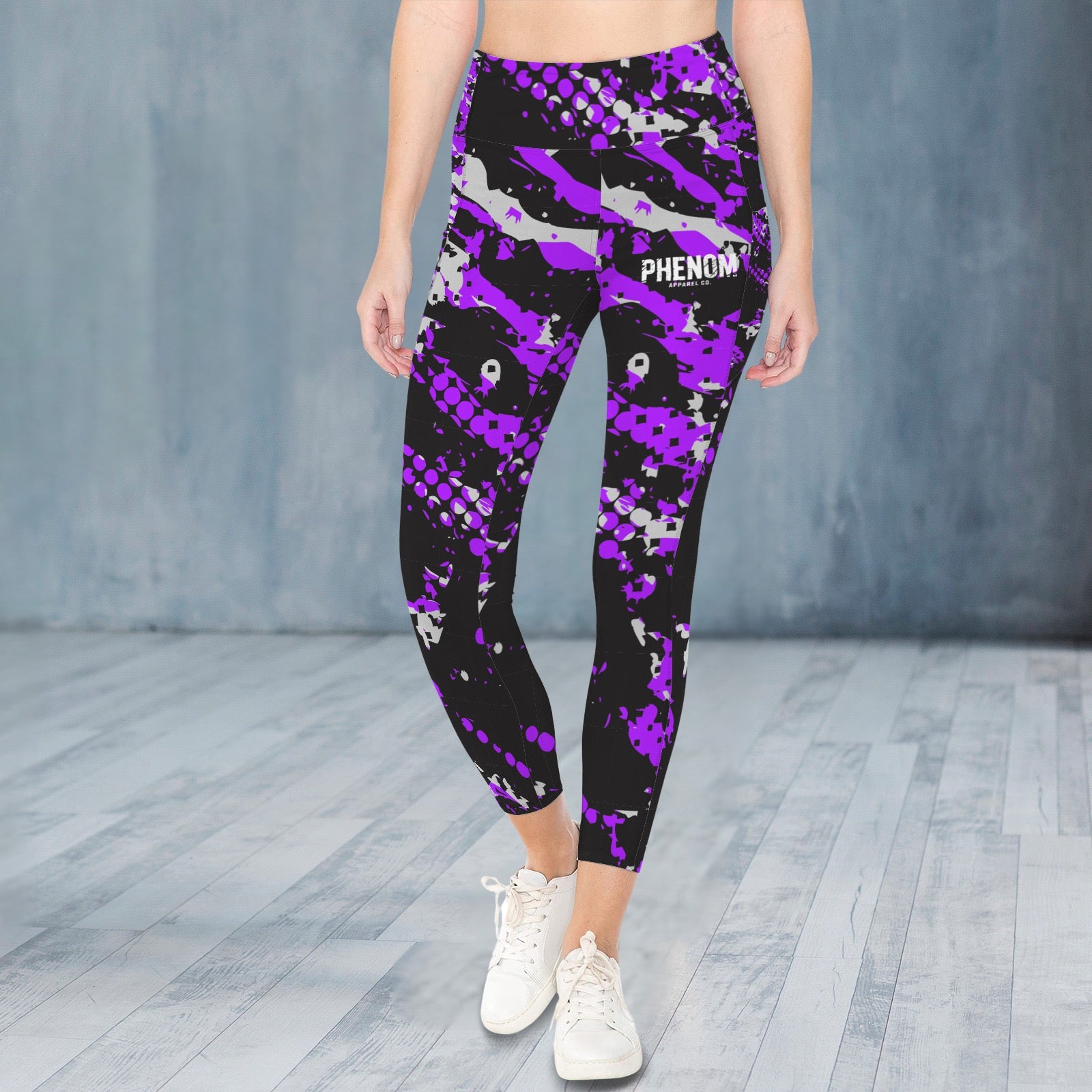 A person modeling high-waisted leggings with a black, purple, and gray camouflage pattern, featuring the word 'PHENOM' on the upper right thigh, paired with white sneakers against a gray studio backdrop.