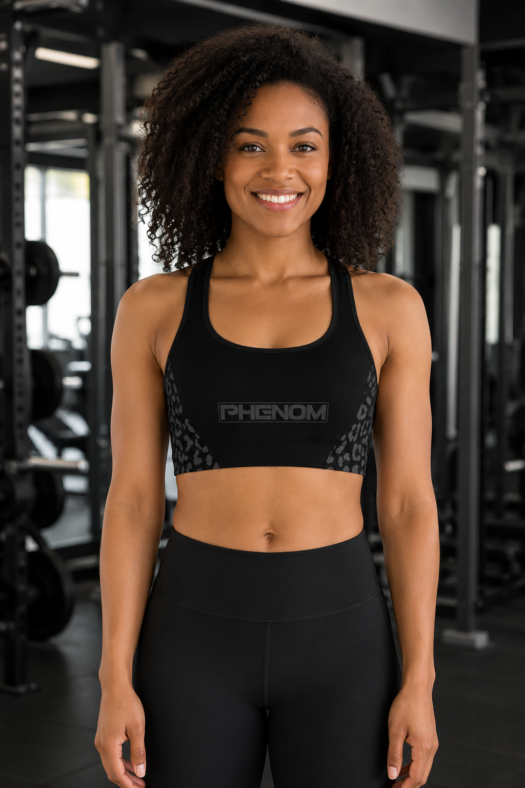 A smiling woman with curly hair stands in a gym wearing a black athletic sports bra with 'PHENOM' printed across the chest and black leggings.